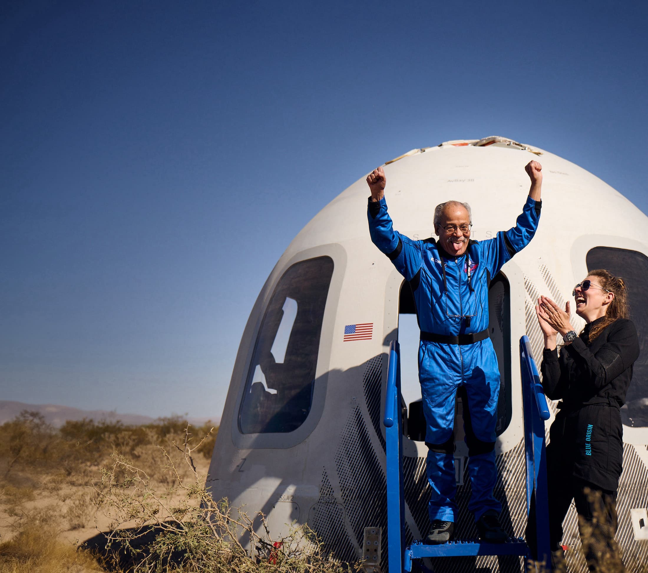 An astronaut in a Blue Origin flight suit steps out of the New Shepard crew capsule with their arms raised above their head.