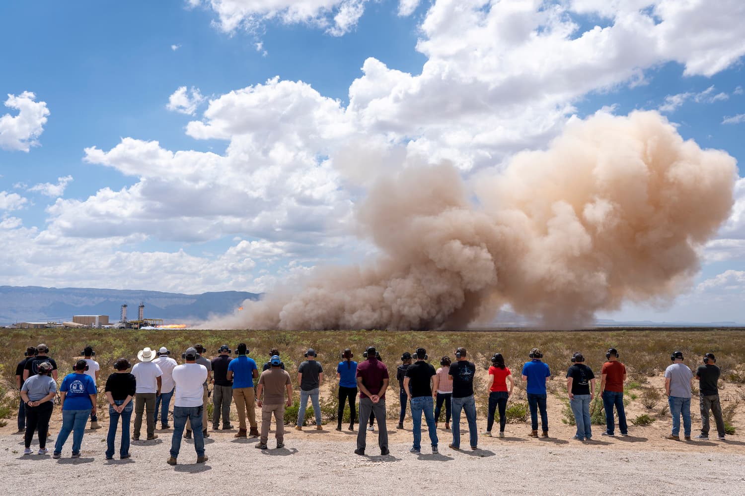 A large group of Blue Origin employees wearing over-ear hearing protection stand with their backs to the camera observing a BE-4 hotfire test from a safe distance away; grey and white exhaust billows in the distance.