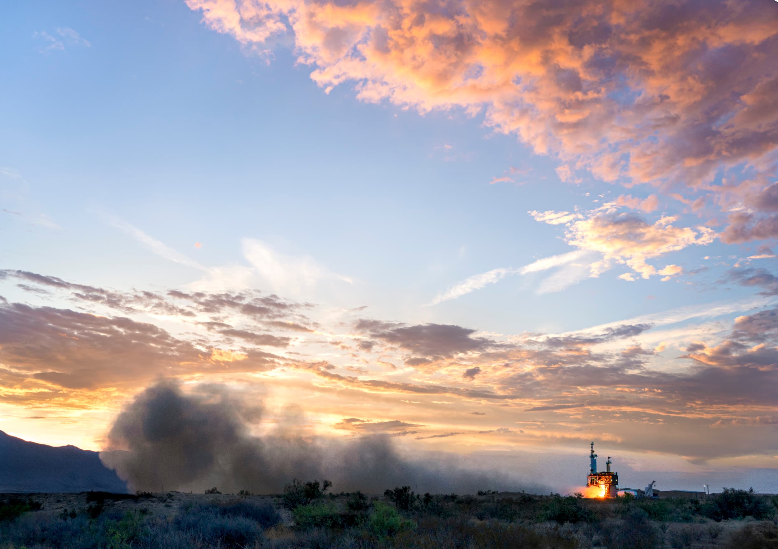 A BE-3U engine is hotfired on the test stand, expelling fiery exhaust against a sky filled with soft orange and purple clouds.