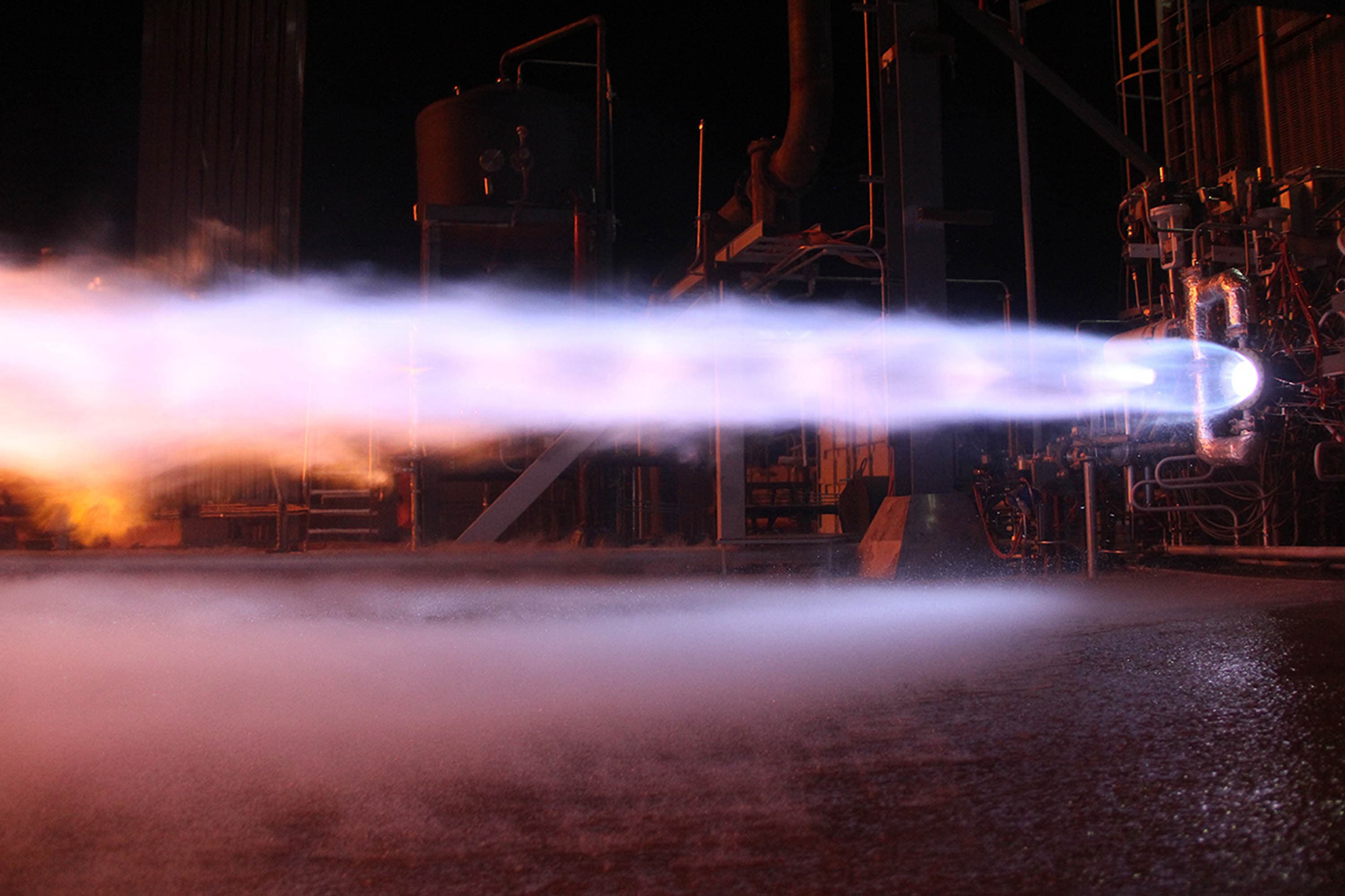 An iridescent, lavender-colored plume of exhaust with mach diamonds expels from an engine mounted on a test stand during a staged-combustion test. The test stand infrastructure is visible in the background.