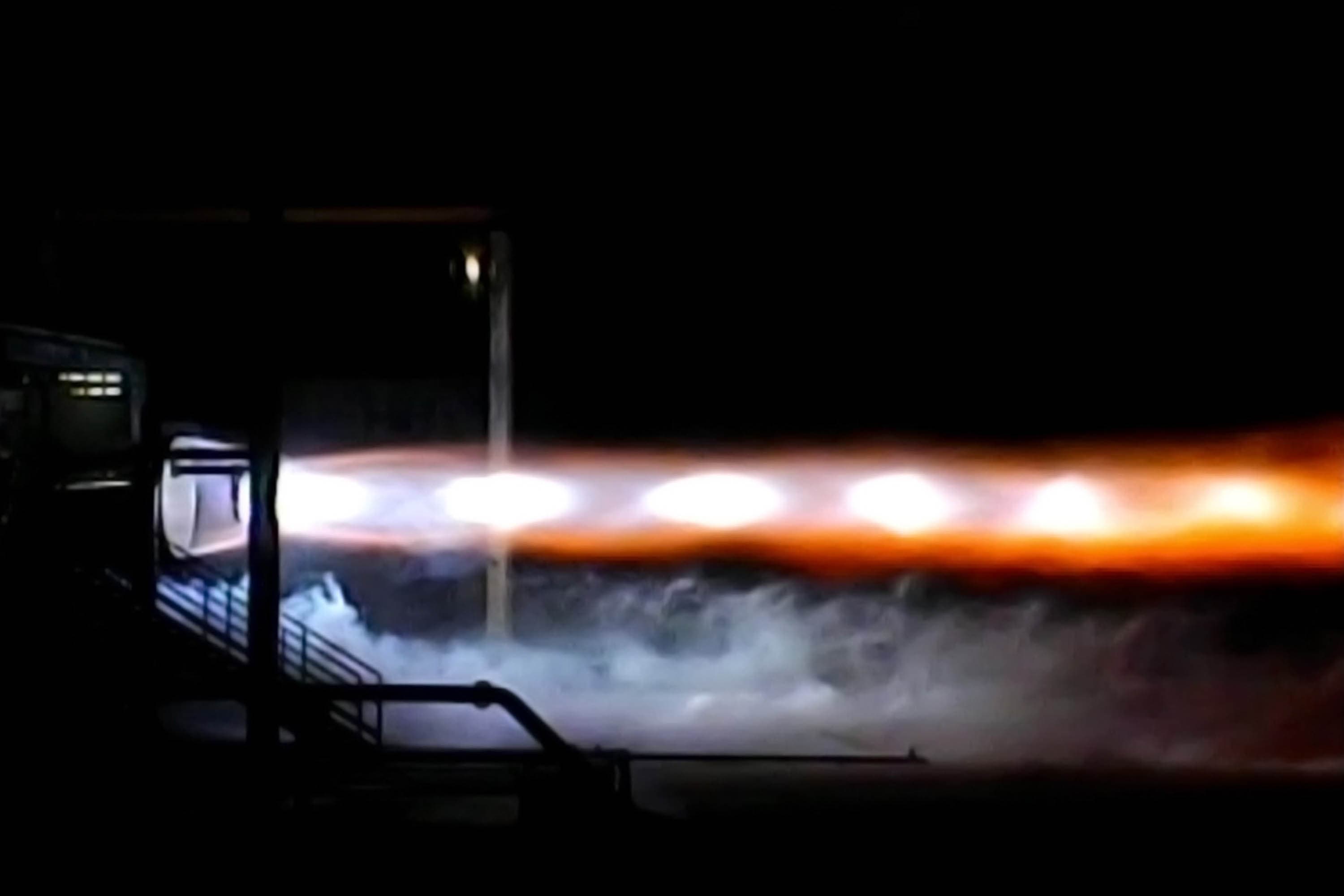 An engine is mounted on the test stand expelling white mach diamonds and a horizontal plume of fiery orange exhaust while wisps of white smoke drift along the ground.