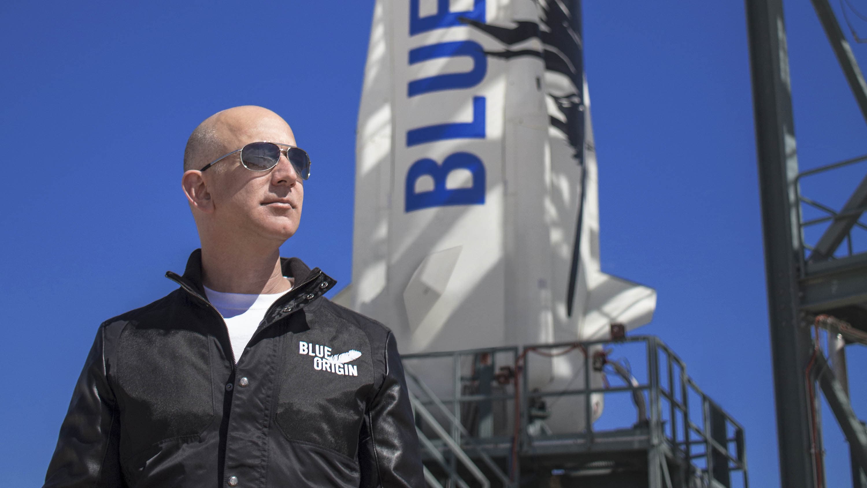 Jeff looks into the distance wearing sunglasses while standing in front of New Shepard perched on the launch tower.
