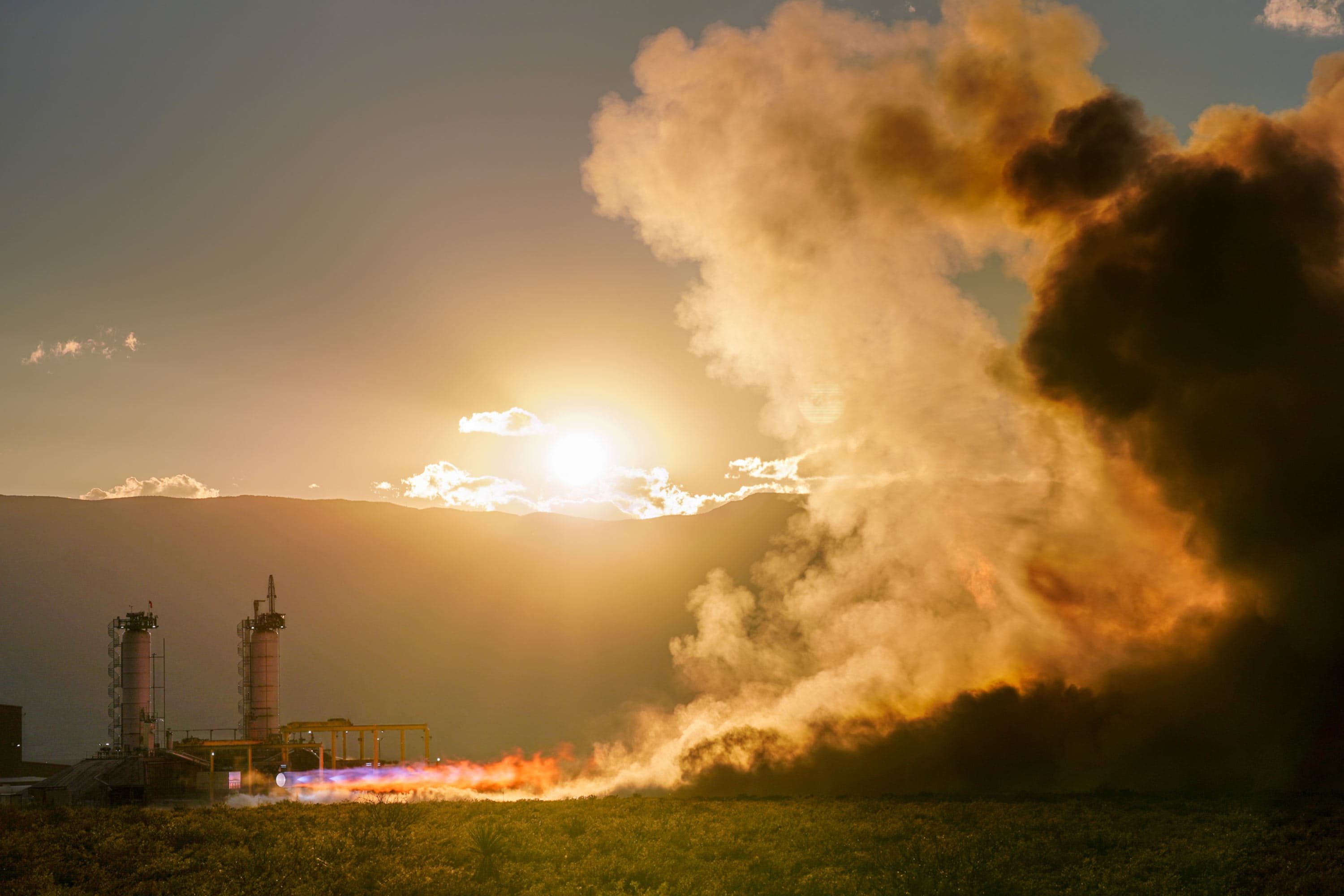 A BE-4 engine is hotfired on the test stand, expelling mach diamonds and fiery exhaust that balloons into large clouds of dust and smoke that fills the right half of the frame. The sun shines brightly over the mountains in the distance.