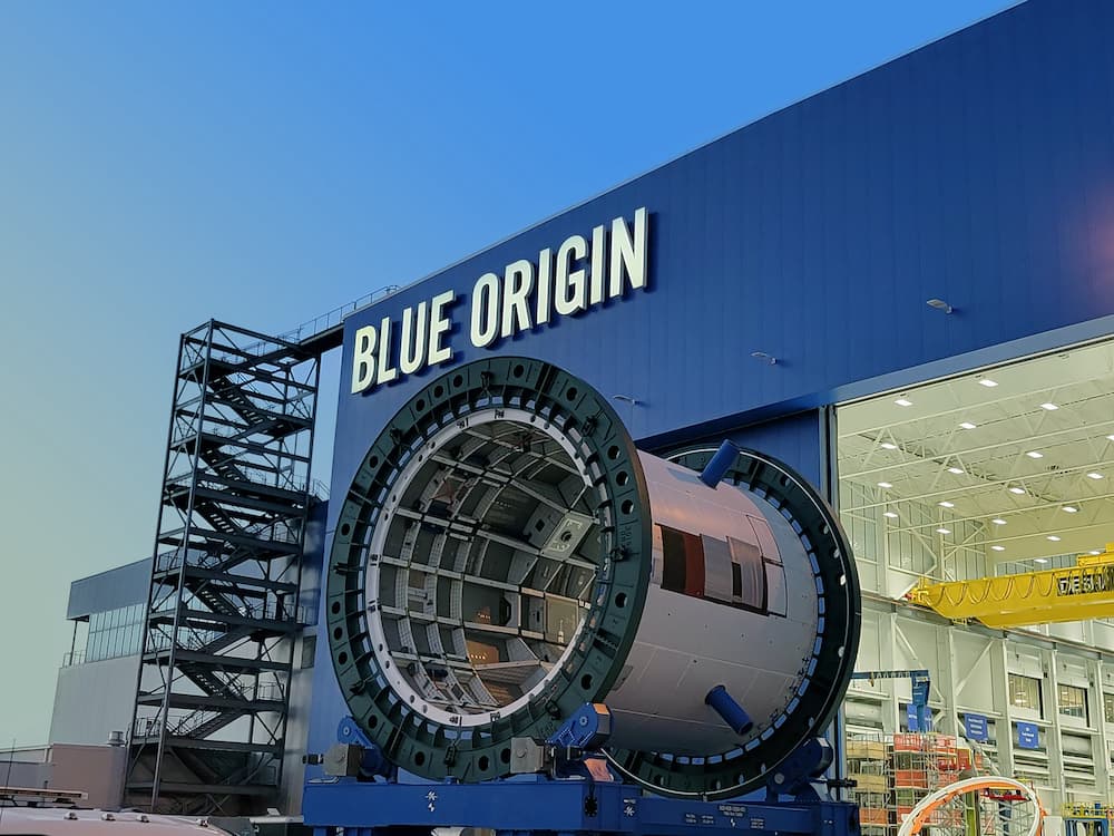 A large cylinder sits on a platform outside a blue building with the words “Blue Origin.” The side of the building is partly open, showing a glimpse of the manufacturing floor inside.