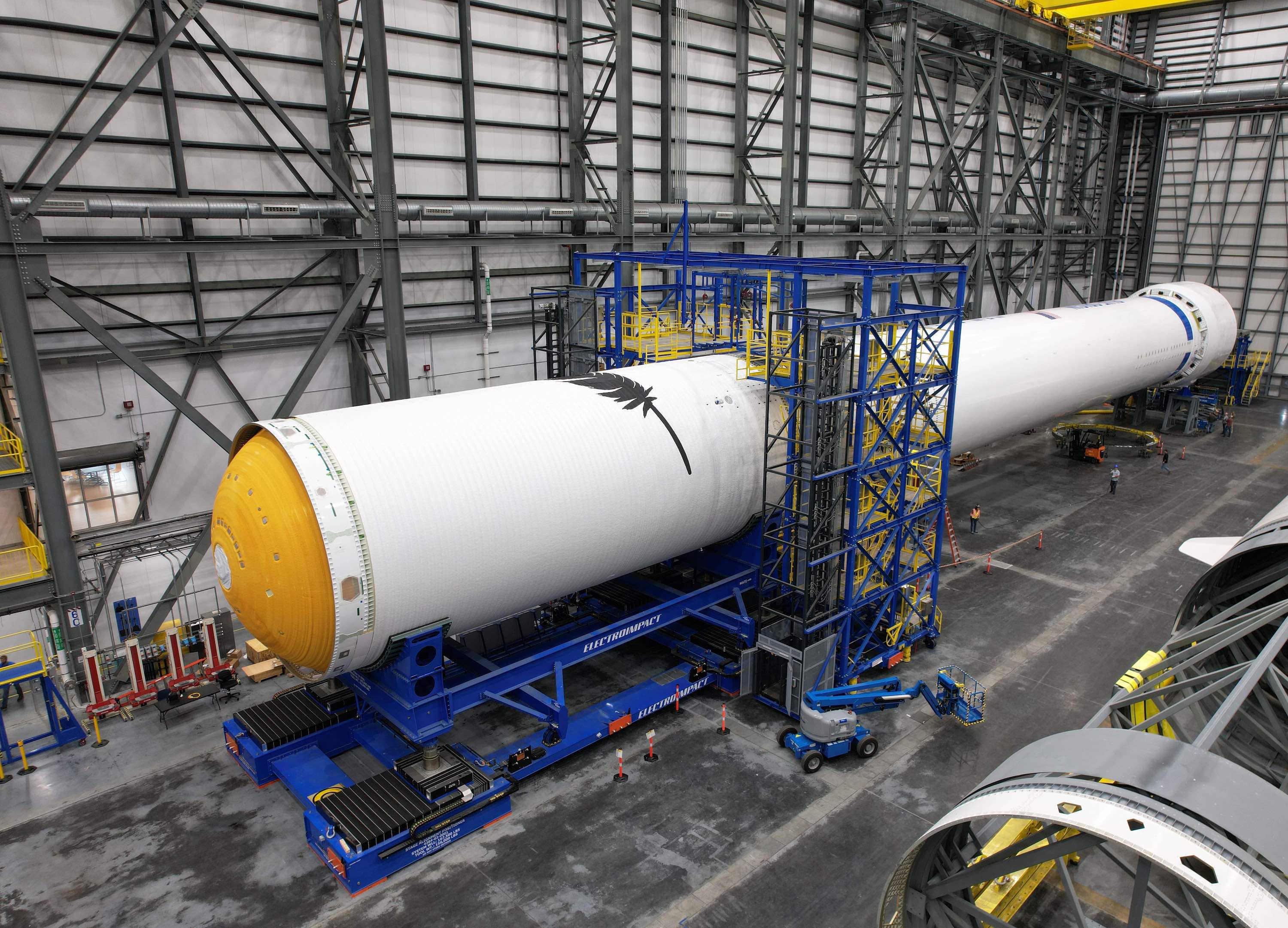 Two long, white cylindrical stages are mated horizontally inside a grey warehouse. Blue scaffolding covers where the two stages were mated. A black Blue Origin feather can be seen on the cylindrical structure.