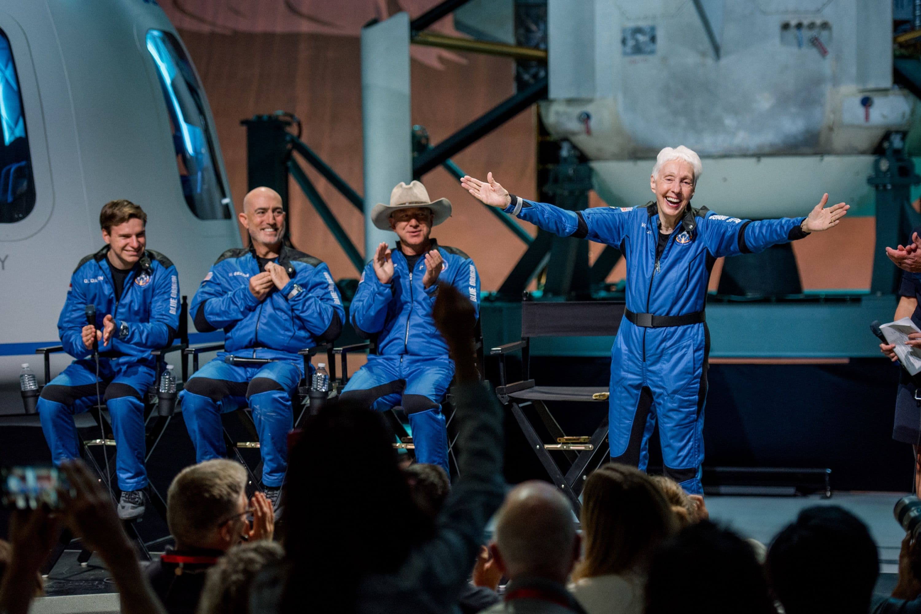 Oliver, Mark and Jeff sit in black directors chairs while Wally stands and smiles with her arms outstretched in celebration.