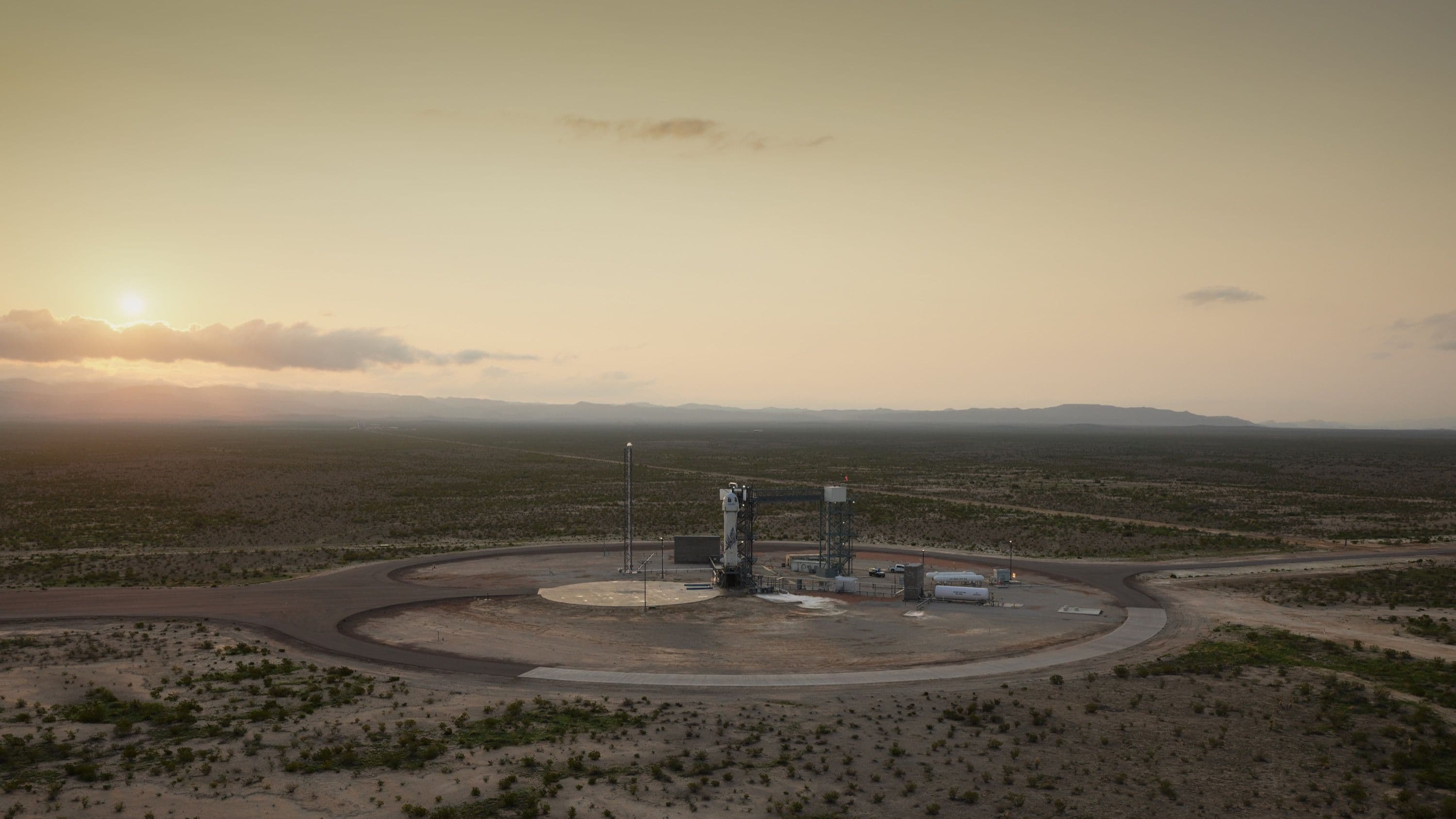 An aerial view at sunrise of New Shepard on the launch pad surrounded by desert and mountains in the distance.
