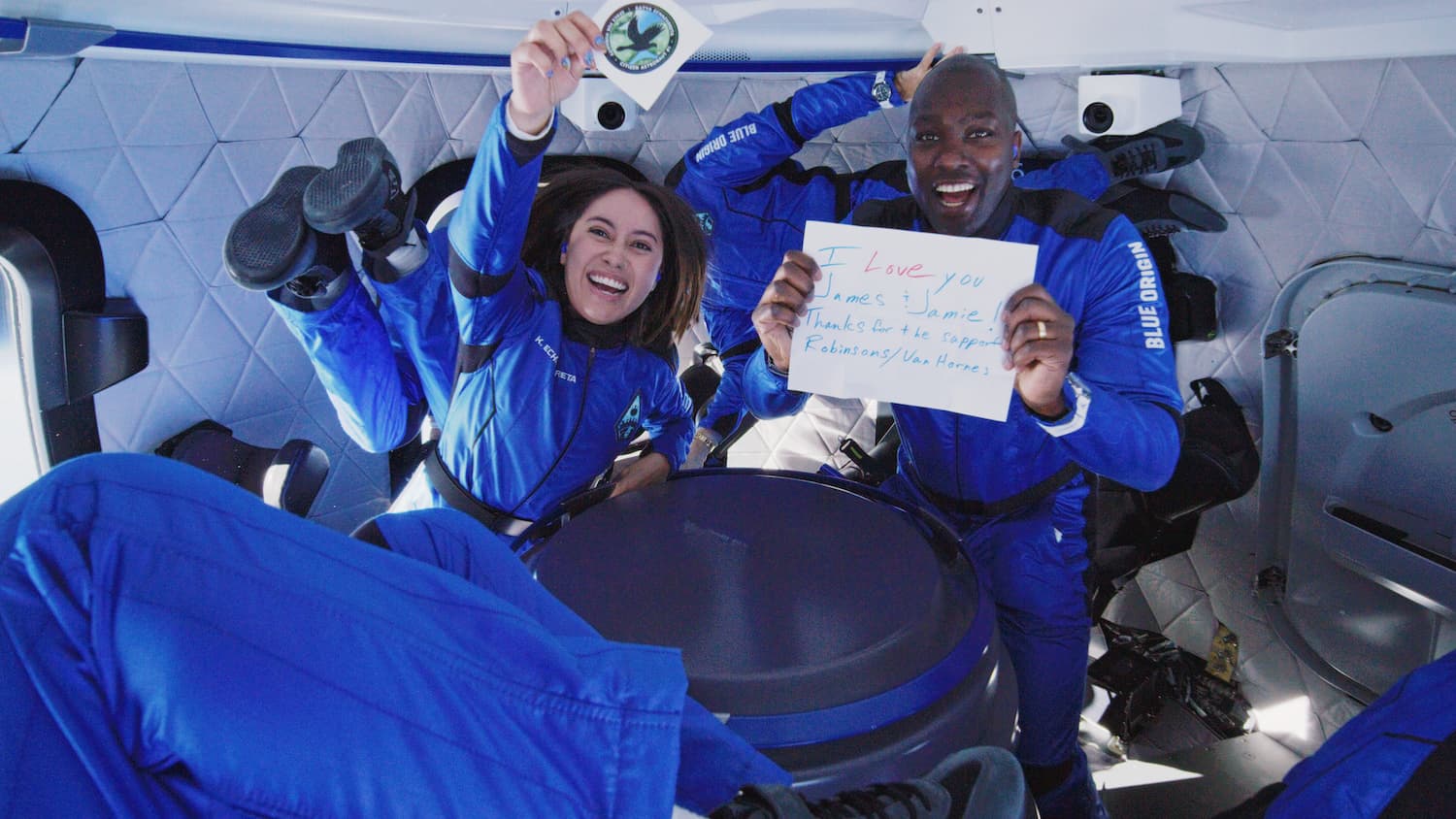 Katya and Jaison float inside the crew capsule. Katya holds a sticker of her Space for Humanity mission patch; Jaison holds a sign that reads "I love you James & Jamie! Thanks for the support Robinsons/Van Hornes."