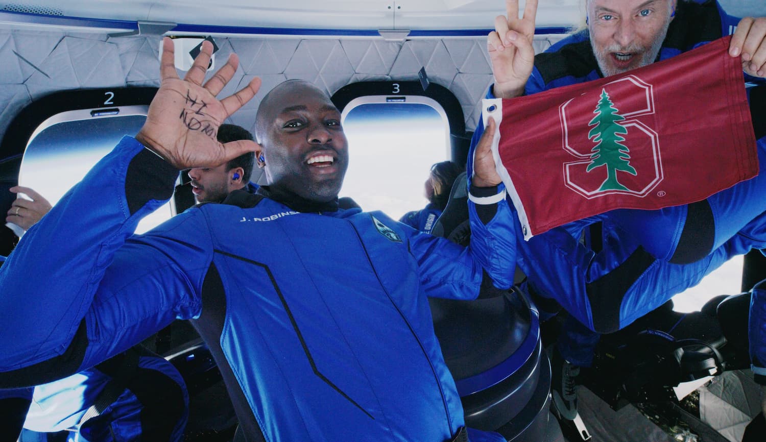 Jaison and Victor float inside the crew capsule. Jaison has "Hi Mom" written on his palm. Victor holds the Stanford flag and makes the peace sign.