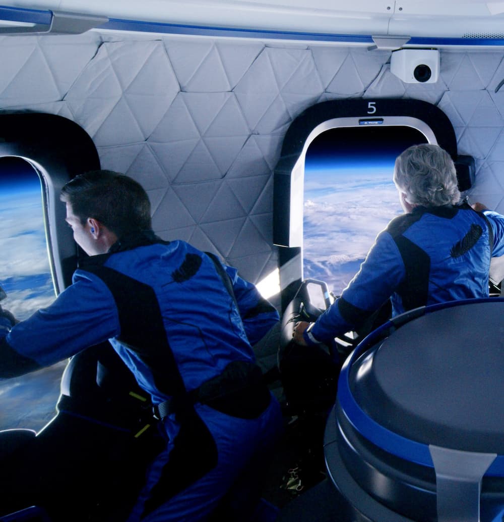 Coby and Steve look out the windows of the crew capsule at the view of Earth and its thin blue atmosphere below them.
