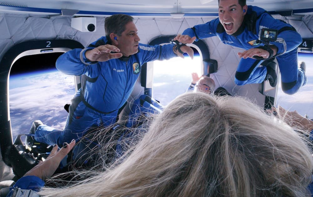 Mario and Coby float inside the crew capsule with their arms raised; a view of Earth and its thin blue atmosphere show through the windows behind them.