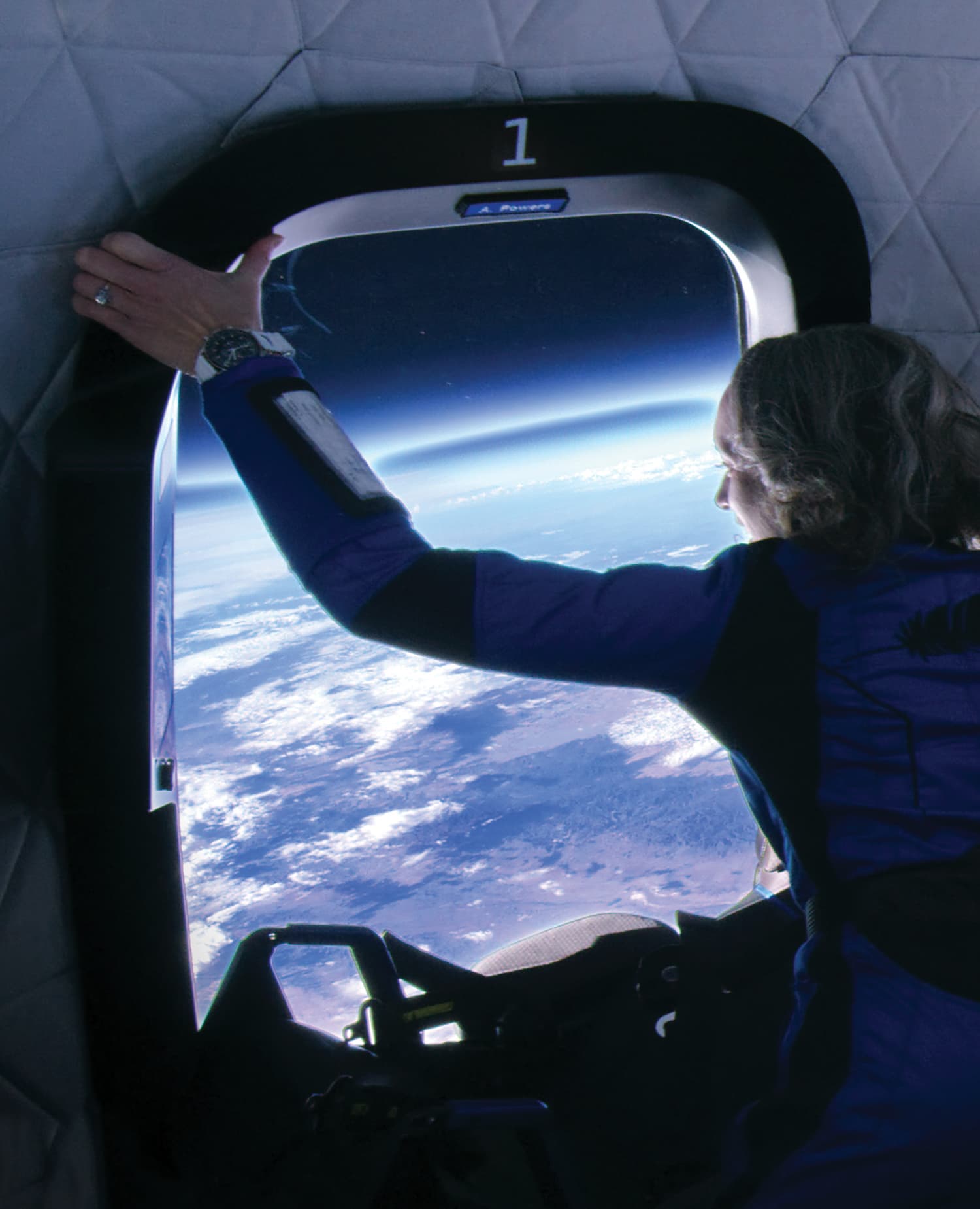 Audrey holds onto the window frame inside the crew capsule, looking at the view of Earth.