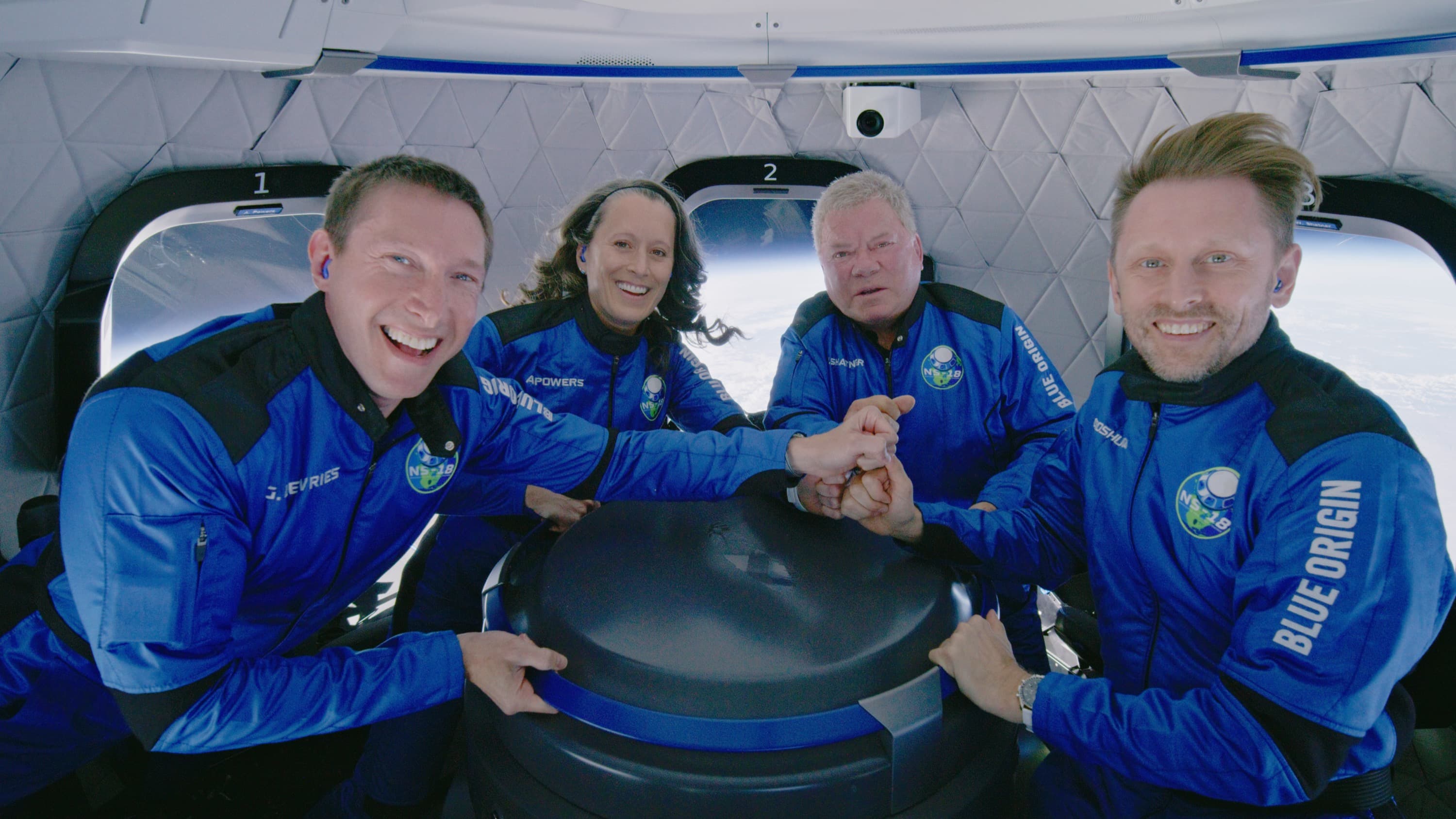 Four smiling astronauts hold their fists on top of each other while posing for a picture and floating inside the crew capsule.