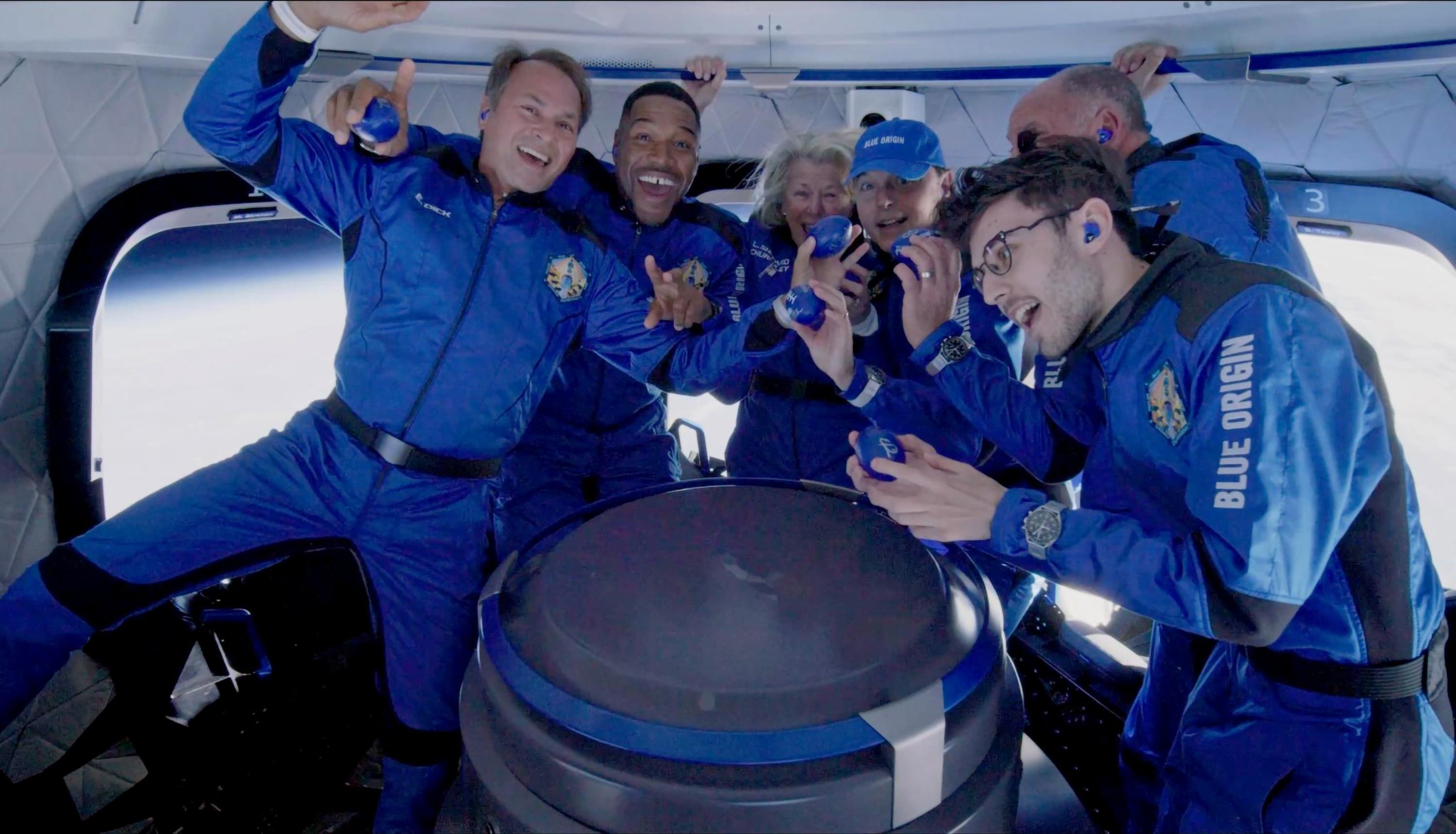Six astronauts of mixed genders and ethnicities smile and pose in a semi-circle around the solid rocket motor as they float inside the crew capsule. Several hold small, blue footballs.