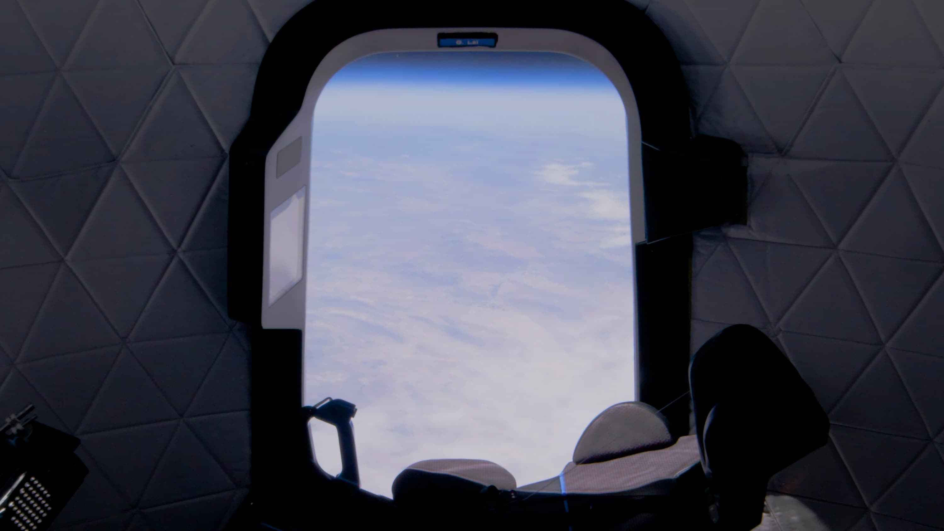 A view of the cloud-covered Earth and thin blue line of its atmosphere from inside the uncrewed crew capsule during flight. The sun reflects on an empty, black seat in front of the window.