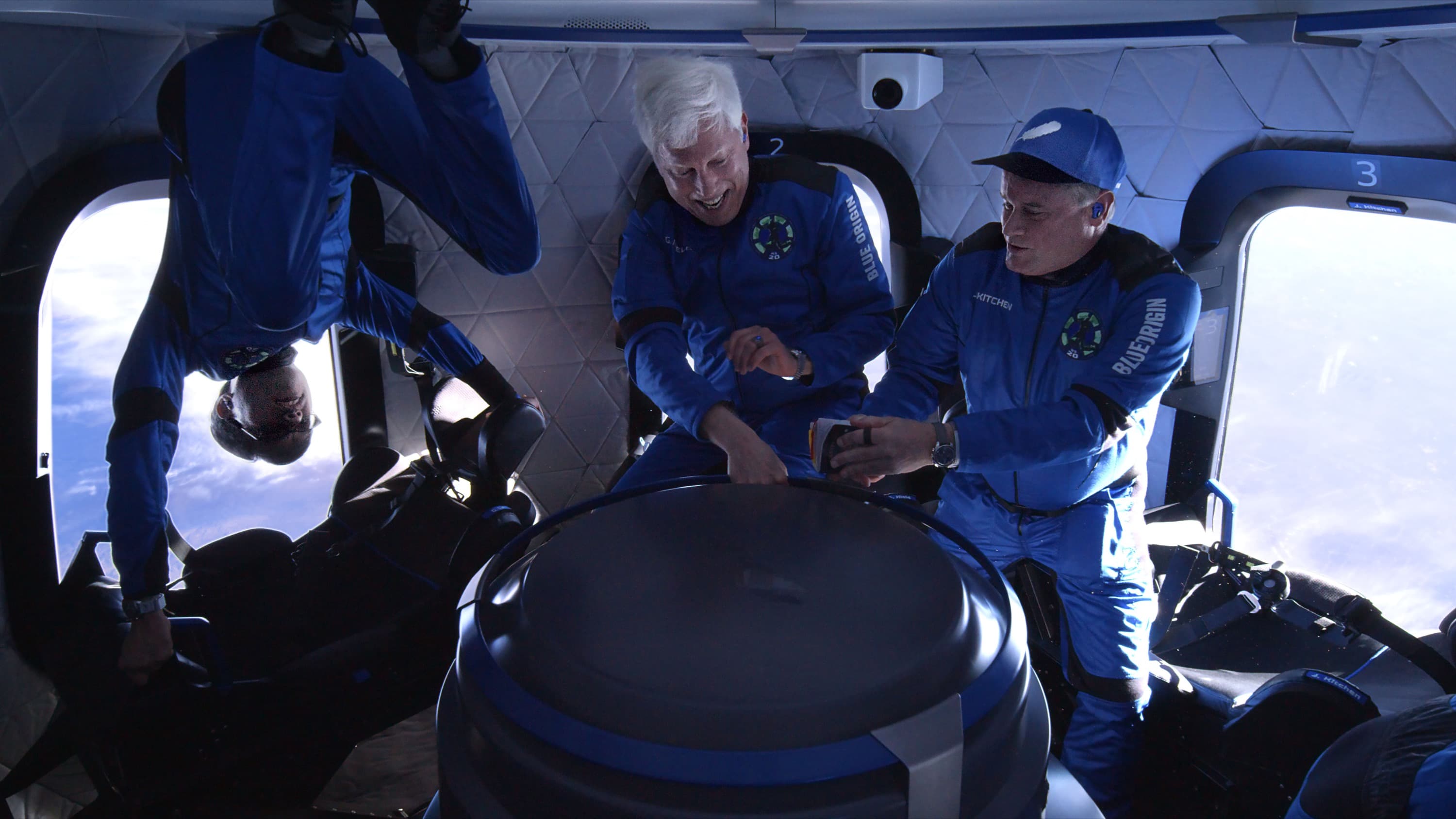 George and Jim float upright while Gary does a flip inside the crew capsule. George smiles while Jim holds his passport.